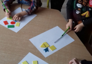 dzieci siedzą przy stole i odrysowują na kartkach figury geometryczne
