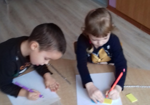 dzieci siedzą przy stole i odrysowują na kartkach figury geometryczne