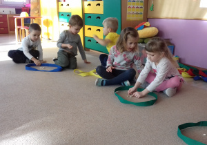 dzieci siedzą na dywanie, mają szarfy i fasolki. Wykonują zadania matematyczne