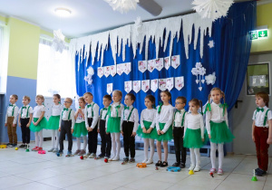 Cała grupa dzieci stoi na scenie podczas występu. Przed nimi na podłodze ułożone są instrumenty. W tle widzimy dekoracje.