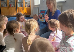 grupa dzieci siedzi na dywanie w bibliotece, słuchają pani bibliotekarki, która pokazuje im książki