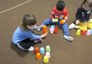 troje dzieci siedzi na dywanie i układa wieże z kolorowych kubeczków