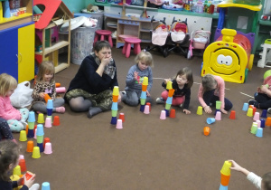 dzieci siedzą w kole na dywanie i układają wieże z kolorowych kubeczków