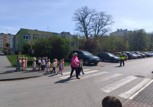 grupa dzieci przechodzi po pasach, jest z nimi nauczycielka, obok stoi policjant