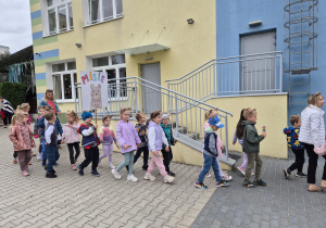 grupa przedszkolaków i ich panie w kolorowym korowodzie wychodzą z przedszkola, jedno z dziećmi niesie plakat z napisem MISIE