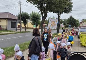 grupa przedszkolaków i ich panie w kolorowym korowodzie wychodzą z przedszkola, jedno z dziećmi niesie plakat z napisem Pszczółki
