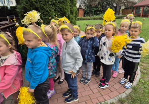 dzieci w przedszkolnym ogrodzie, niektóre mają na głowach opaski z pszczółkami , inne trzymaja w rękach żółte pompony z bibuły