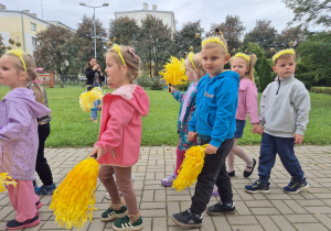 dzieci w przedszkolnym ogrodzie, niektóre mają na głowach opaski z pszczółkami , inne trzymaja w rękach żółte pompony z bibuły