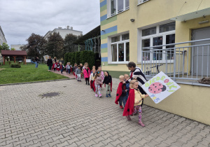 grupa przedszkolaków i ich panie w kolorowym korowodzie wychodzą z przedszkola, jedno z dziećmi niesie plakat z napisem MISIE , inne z napisem BIEDRONKI
