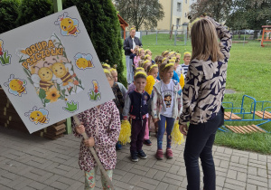 nauczycielka i dzieci (z opaskami w pszczólki) stoją w ogrodzie przedszkolnym, jedno z nich na plakat z napisem Pszczółki
