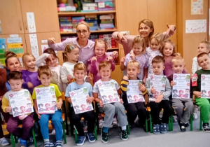 Grupa dzieci z nauczycielkami pozują do zdjęcia. Chłopcy siedzą na krzesełkach i prezentują dyplomy.