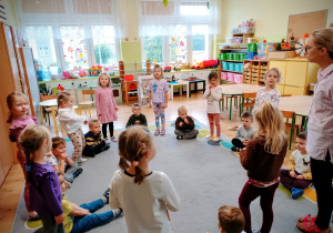 Chłopcy siedzą na dywanie w kole. Pomiędzy nimi stoją dziewczynki. Na dalszym planie widać nauczycielkę.