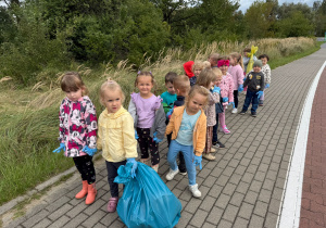 grupa dzieci idzie chodnikiem, mają na rękach jednorazowe rękawiczki, jedno z dziecmi trzyma niebieski worek na odpady, inne zółty
