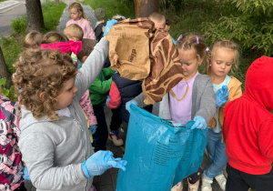 grupa dzieci , jedne stoją na chodniku, inne na łące, zbierają śmieci , jedno dziecko trzyma worek na odpady
