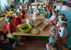 dzieci w opaskach z obrazkiem jabłka siedzą przy stoliku na którym połozone są produkty na pieczenie ciasta