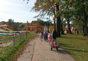 Dzieci spacerują w parach. Nauczycielka idzie za dziećmi i ciągnie przyczepkę z ozdobionymi dyniami.