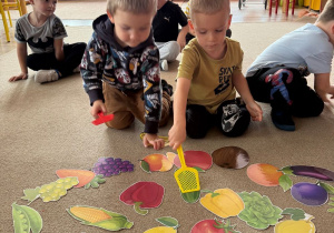 na dywanie leżą kolorowe obrazki owoców i warzyw, przy nich siedządzieci, które mają w rękach łapki na muchy, którymi uderzająw obrazki, na drukm planie pozostałe dzieci