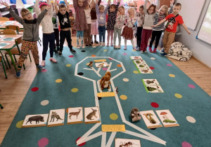 grupa dzieci stoi na dywanie w klasie i unosi ręce, na dywanie naklejony schemat drzewa, obok liście i owoce drzew, maskotka wiewiórki