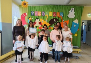grupa pasowanych dzieci, rodziców i nauczycielek pozuje do wspólnego zdjęcia, w tle dekoracja