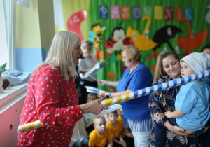 pani dyrektor "pasuje" niebieską kredką przedszkolaka, którego na rękach trzyma mama , w tle inne osoby i dekoracja