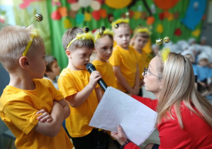 na pierwszym planie grupa dzieci przebranych za pszczółki , przy nich nauczycielka, która jednemu z dzieci przytrzymuje mikrofon, w tle dzieci z pozostałych