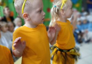 dwoje dzieci przebranych za pszczólki, pokazują treść piosenki, którą śpiewają, w tle inne dzieci