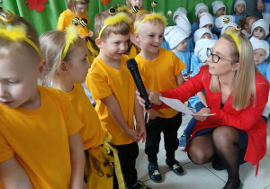 na pierwszym planie grupa dzieci przebranych za pszczółki , przy nich nauczycielka, która jednemu z dzieci przytrzymuje mikrofon, w tle dzieci z pozostałych