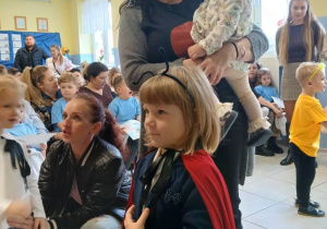 dziewczynka przebrana za biedronkę przygotowuje siędo pasowania, obok stoi jej mama z dzieckiem na ręce, w tle inne osoby