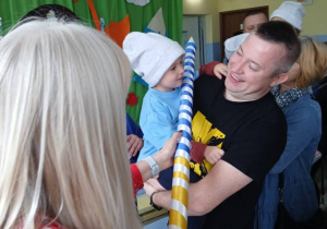 pani dyrektor "pasuje" niebieską kredką przedszkolaka, którego na rękach trzyma tata , w tle inne osoby i dekoracja