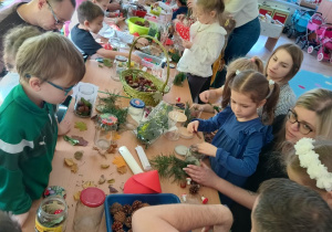 dzieci i rodzice tworzą z darów jesienie kompozycje w śłoikach. siedzą przy stołach z rozłożonymi matariałami do pracy