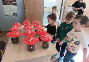 grupa 4 dzieci ogląda wykonane na warsztatach prace (słoiki z darami lasu nakryte kartonikiem imitującym muchomory) , które leżą na stole