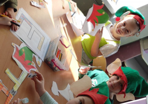 dzieci w czapkach elfów pracują przy stoliku, na nim leżą kolorowanki z literą E