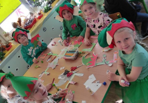 dzieci w czapkach elfów pracyją przy stoliku, na nim leżą wycięte kolorowanki z literą E