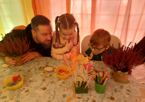 rodzic i dwoje przedszkolaków, siedzą przy stole na którym leżą wrzosy, mazaki i ozdobne listki