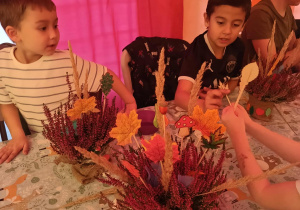 troje przedszkolaków, siedzą przy stole na którym leżą wrzosy, mazaki i ozdobne listki