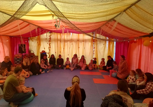 grupa dzieci i rodziców w ogromnym namiocie, wszyscy siedzą w kole, wokół tiulowe dekoracje w kolorach jesieni