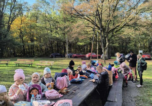 Dzieci siedzą przy długim stole, który znajduje się na łonie natury i zjadają przekąski.