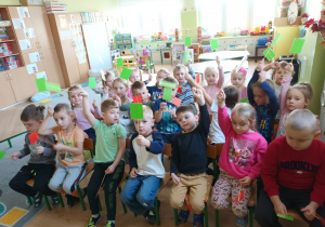 Dzieci siedzą na krzesełkach i podnoszą zielone oraz czerwone karteczki, odpowiadając na zadane pytanie, związane z tematem.