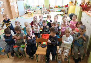 Dzieci siedzą na krzesełkach i prezentują gesty związane z umiejętnością reagowania na sytuacje niebezpieczne.