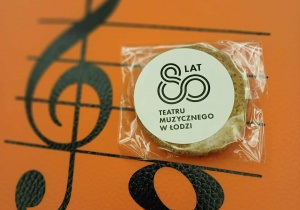 ciastko z napisem 80 lat teatru muzycznego leży na pomarańczowej podkładce z kluczem wiolunowym