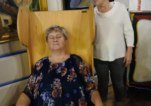 "Gniazdo" w Felicjanowie. Dwie panie, jedna siedzi w drewniamy fotelu ze strunami , druga stoi za nią i "gra"