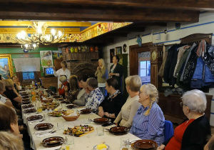 wnętrze starej chaty, przy stole siedzą uczestnicy wyjazdu do "Gniazda", słuchająhistorii tego miejsca opowoadanej przez właścicielkę