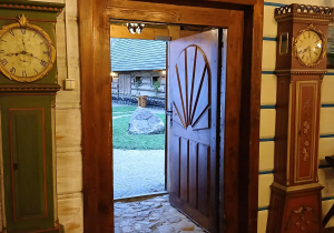 wnętrze jednej z chat w "Gnieździe", otwarte drewniane drzwi, widać podwórko, obok wejścia stary zegar