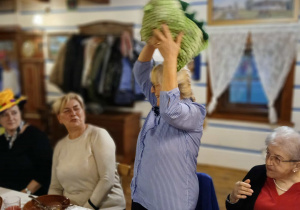 uczestnicy spotkania w Gnieżdzie w Felicjanowie biorą udział w aktywnościach teatralnych, kilka osób siedzi przy stole, jedna stoi, na głowie ma śmieszne nakrycie