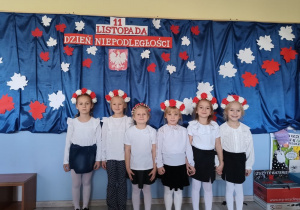 sześć dziewczynek ubranych na galowo, z biało-czerwonymi wiankami na głowach stoi na tle dekoracji z okazji święta niepodległości, pozują do zdjęcia