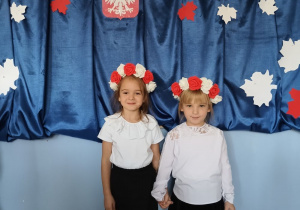 dwie dziewczynki ubrane na galowo, z biało-czerwonymi wiankami na głowach stoi na tle dekoracji z okazji święta niepodległości, pozują do zdjęcia