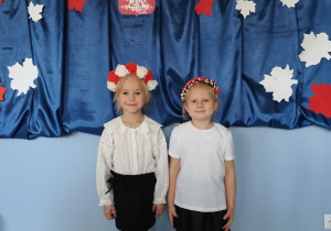 dwie dziewczynki ubrane na galowo, z biało-czerwonymi wiankami na głowach stoi na tle dekoracji z okazji święta niepodległości, pozują do zdjęcia