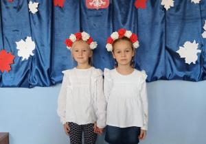 dwie dziewczynki ubrane na galowo, z biało-czerwonymi wiankami na głowach stoi na tle dekoracji z okazji święta niepodległości, pozują do zdjęcia