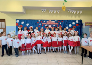 grupa dzieci ubranych na galowo, z biało-czerwonymi wiankami na głowach stoi na tle dekoracji z okazji święta niepodległości, pozują do zdjęcia