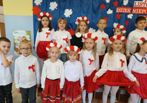 grupa dzieci ubranych na galowo, z biało-czerwonymi wiankami na głowach stoi na tle dekoracji z okazji święta niepodległości, pozują do zdjęcia